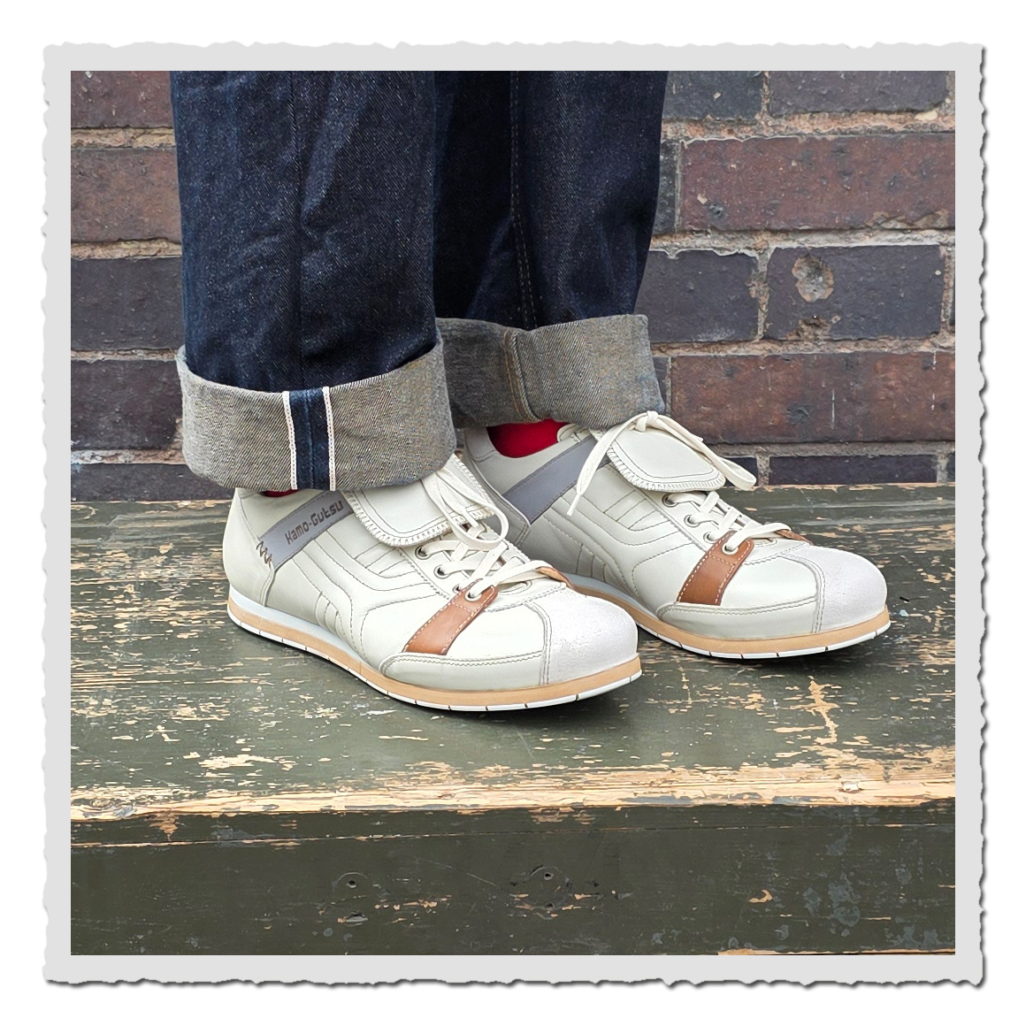 Altweiße Schnürschuhe mit heller Lasche an einer Person mit dunklen Jeans vor Backsteinwand auf grünem Holzpodest.